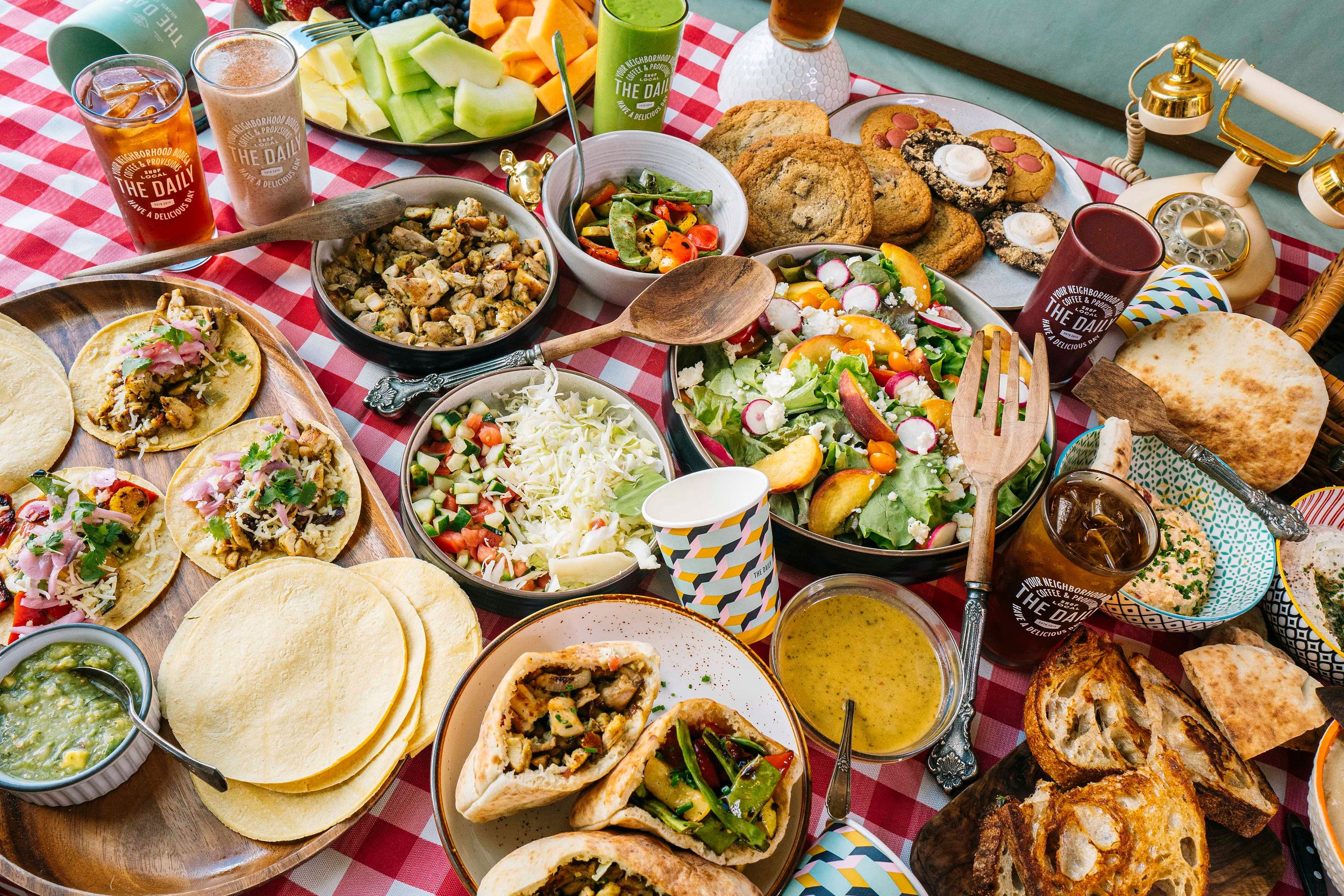 A colorful spread of breakfast and brunch foods on a red checkered tablecloth, including fresh fruit, salads, tacos, peppers, bread, pastries, and beverages for catering