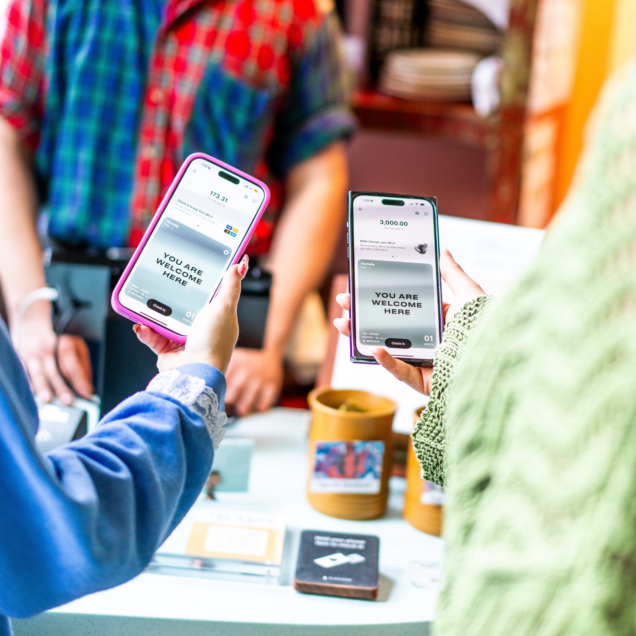 Two people holding smartphones displaying welcome screens at a reception desk, with a person behind the counter and various items on the table in the background.