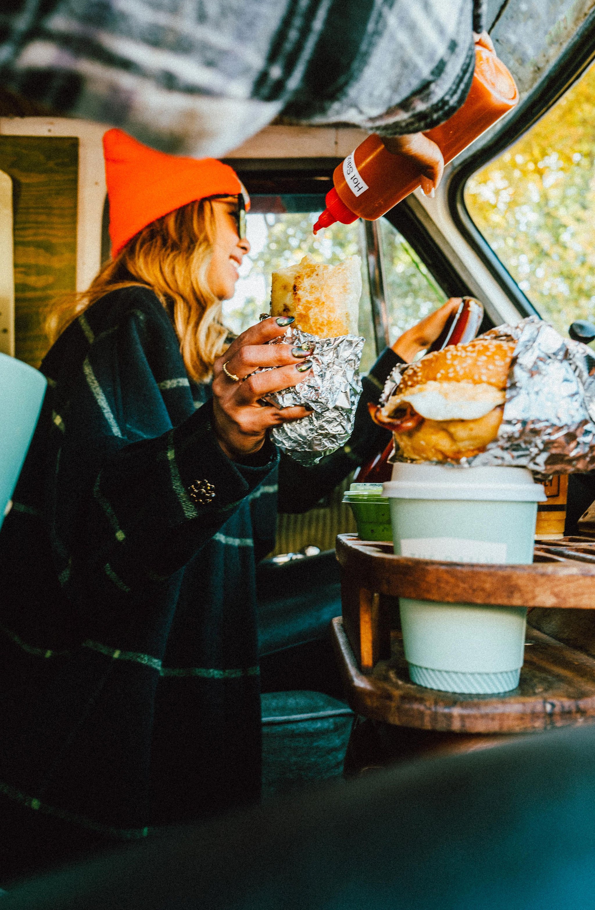Woman with long blonde hair, red beanie, and glasses, smiling and holding a sandwich wrapped in foil inside a food truck amid vibrant, cozy autumn setting.