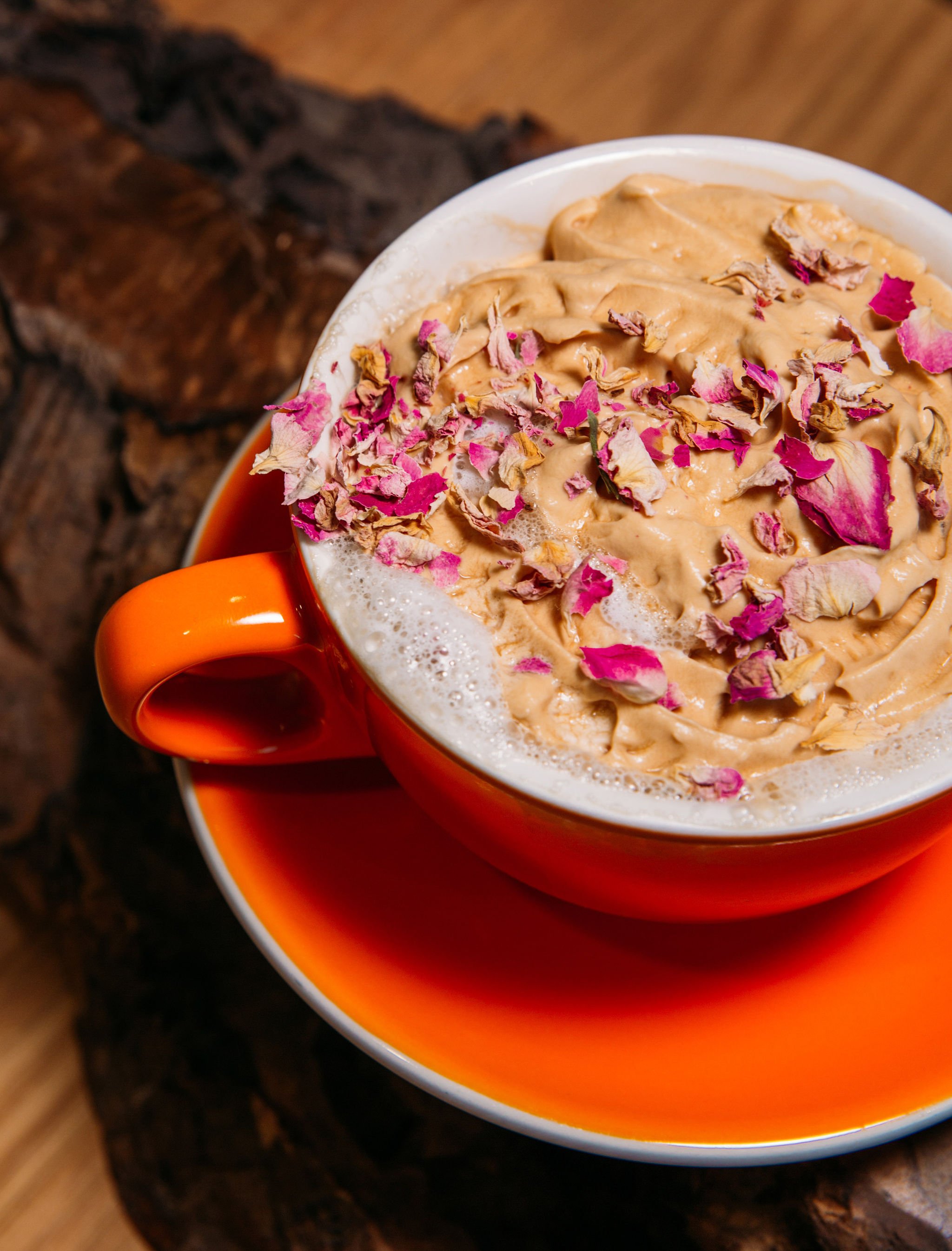 A close-up of a orange mug filled with a frothy beverage topped with whipped cream, pink dried rose petals, and a hint of chocolate shavings, placed on a matching orange saucer on a wooden surface.