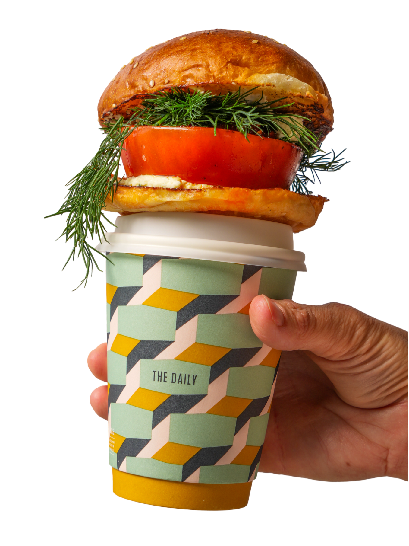 Hand holding a disposable coffee cup that contains a sandwich with a bun, tomato, herbs, egg, and lettuce, with the sandwich items spilling over the top.